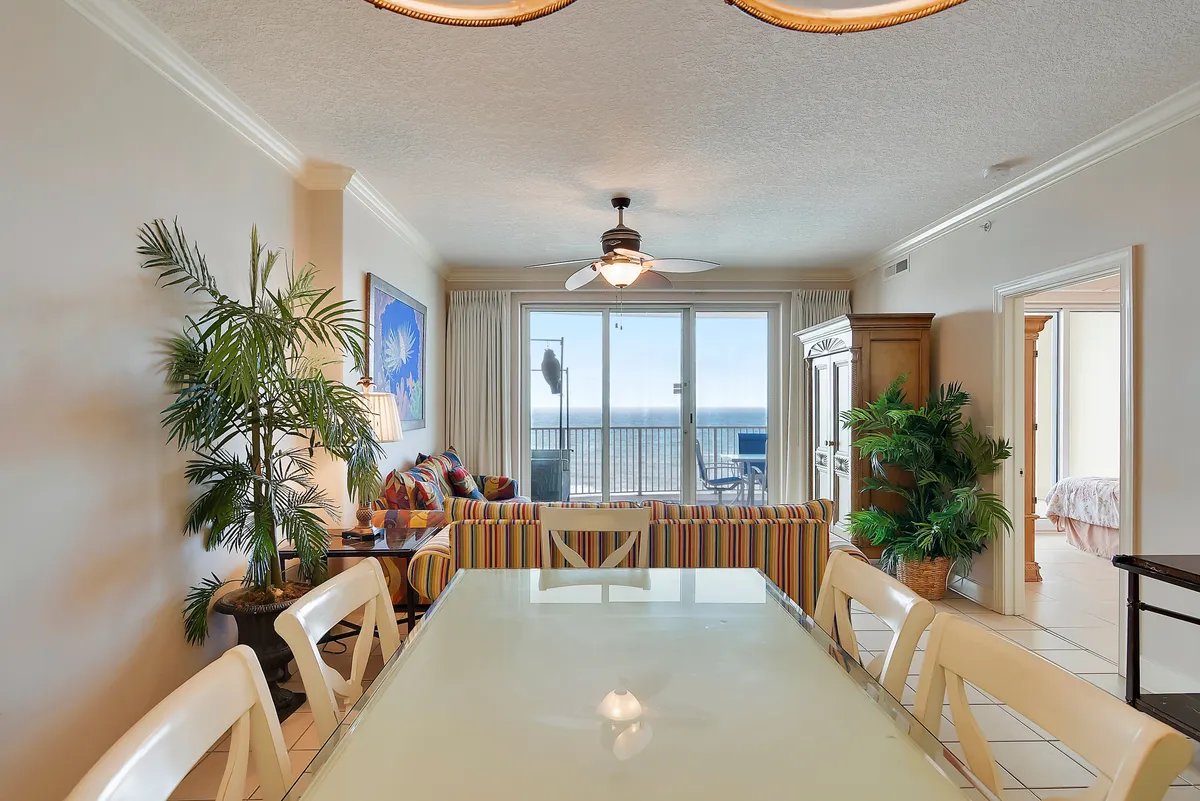 This image showcases a sunlit open-plan living and dining area with a colorful sofa, dining table, potted plants, and large windows that open to stunning ocean views. The space is bright, welcoming, and features contemporary décor, perfect for gatherings or relaxing moments.