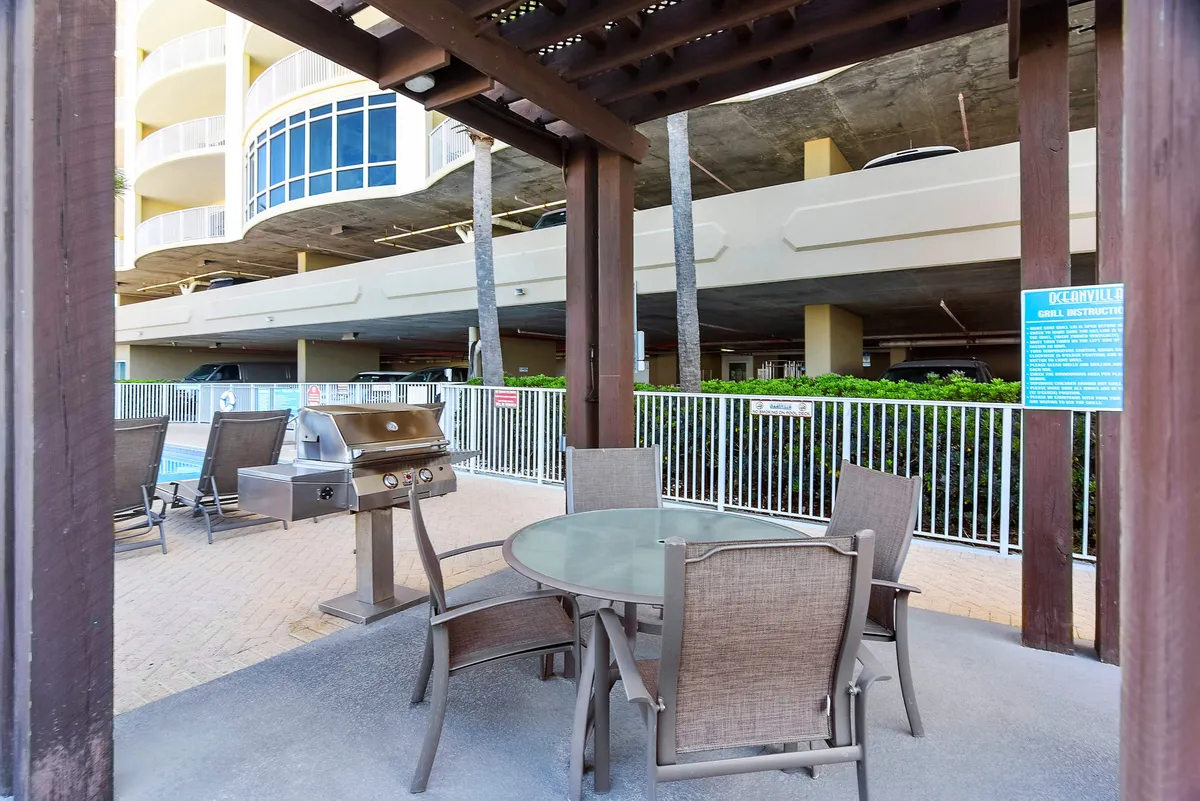 An inviting BBQ pavilion area with a grill, table, and chairs situated next to a pool and surrounded by palm trees, creating a perfect setting for outdoor group dining and gatherings. The vibrant atmosphere encourages social interaction and relaxation for families and friends.