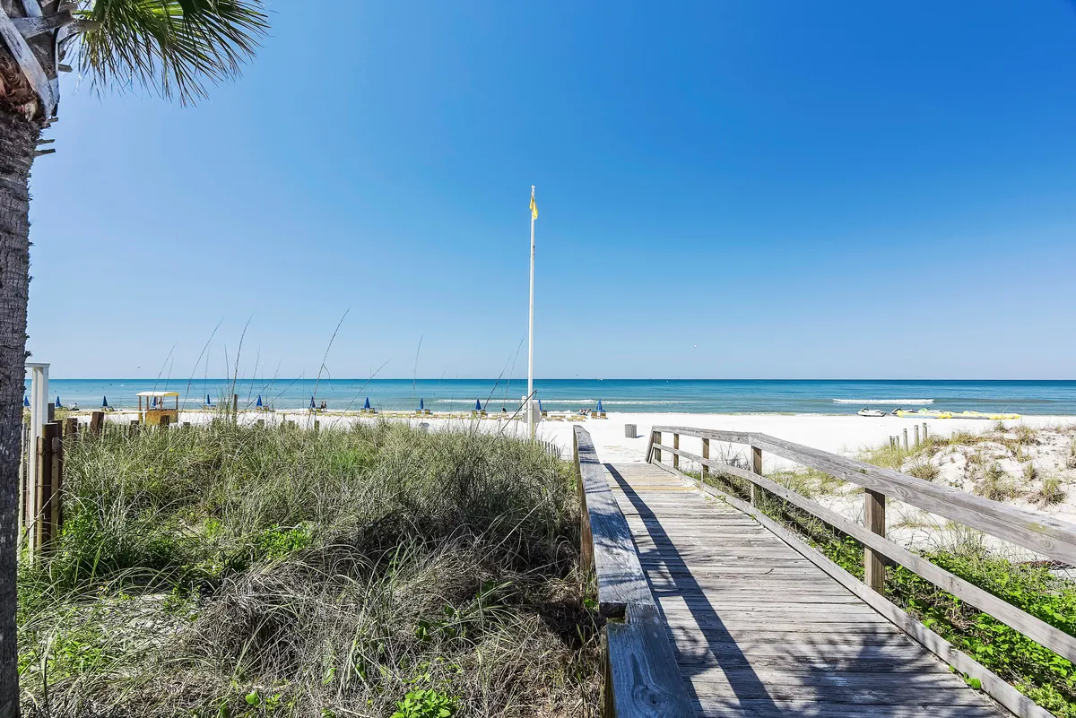 A picturesque wooden walkway leads directly to the sandy beach and ocean, framed by a palm tree and blue sky, evoking the allure of private strolls and easy beach access.