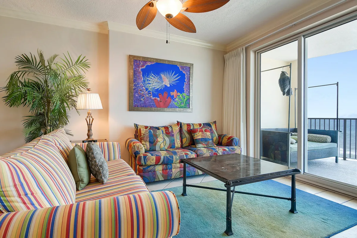 A vibrant living room featuring colorful patterned couches, a floor-to-ceiling sliding glass door leading to the balcony, large ocean-themed artwork, and a blue gradient area rug. The space is filled with natural light, offering a seamless visual connection to the balcony and Gulf views beyond.