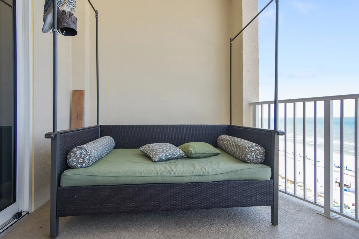 A serene private balcony features a wicker daybed with green cushions and patterned bolsters, set against a stunning backdrop of the sandy beach and expansive ocean. The blue sky and endless horizon enhance the tranquil, rejuvenating atmosphere, inviting guests to relax, read, or sip a drink while enjoying the fresh sea breeze.