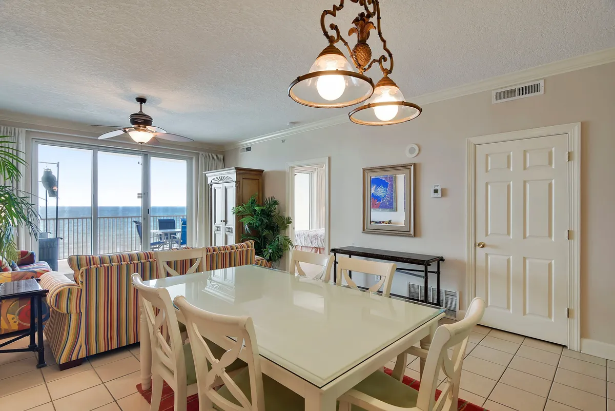 An airy, open-plan living and dining area with a large dining table and white chairs, striped sofa facing the ocean, and expansive floor-to-ceiling windows overlooking the beach and balcony. A decorative light fixture and coastal accents create a sense of casual elegance and comfort.