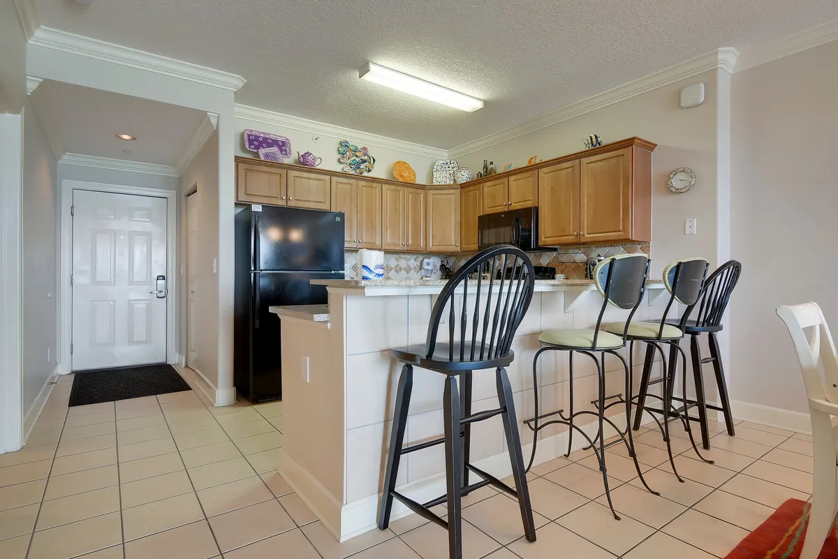 A modern kitchen featuring a central island with three bar stools, black refrigerator, wooden cabinetry, and an open entryway. The island invites social interaction and casual dining, while the layout visually connects the kitchen to the rest of the living space.