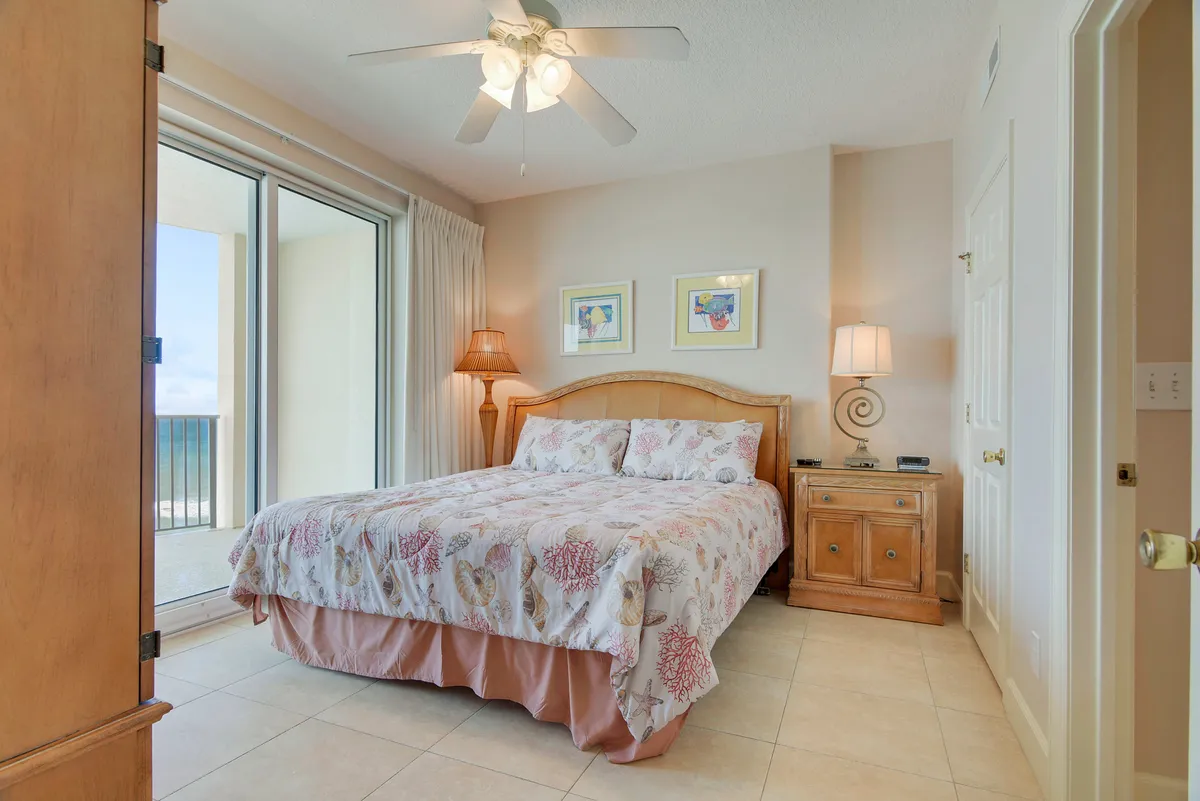 This image features a serene master bedroom with a king-sized bed adorned in seashell-themed bedding. Floor-to-ceiling sliding glass doors open directly onto a private balcony with ocean views, and two bedside tables with lamps add a cozy ambiance. A ceiling fan with a light fixture is visible above.