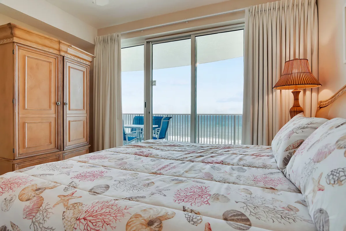 A peaceful, spacious bedroom featuring a king-sized bed with coral and seashell bedding. Large wooden armoire and a bedside lamp add to the inviting decor. Sliding glass doors provide a glimpse of the private balcony and breathtaking gulf views.
