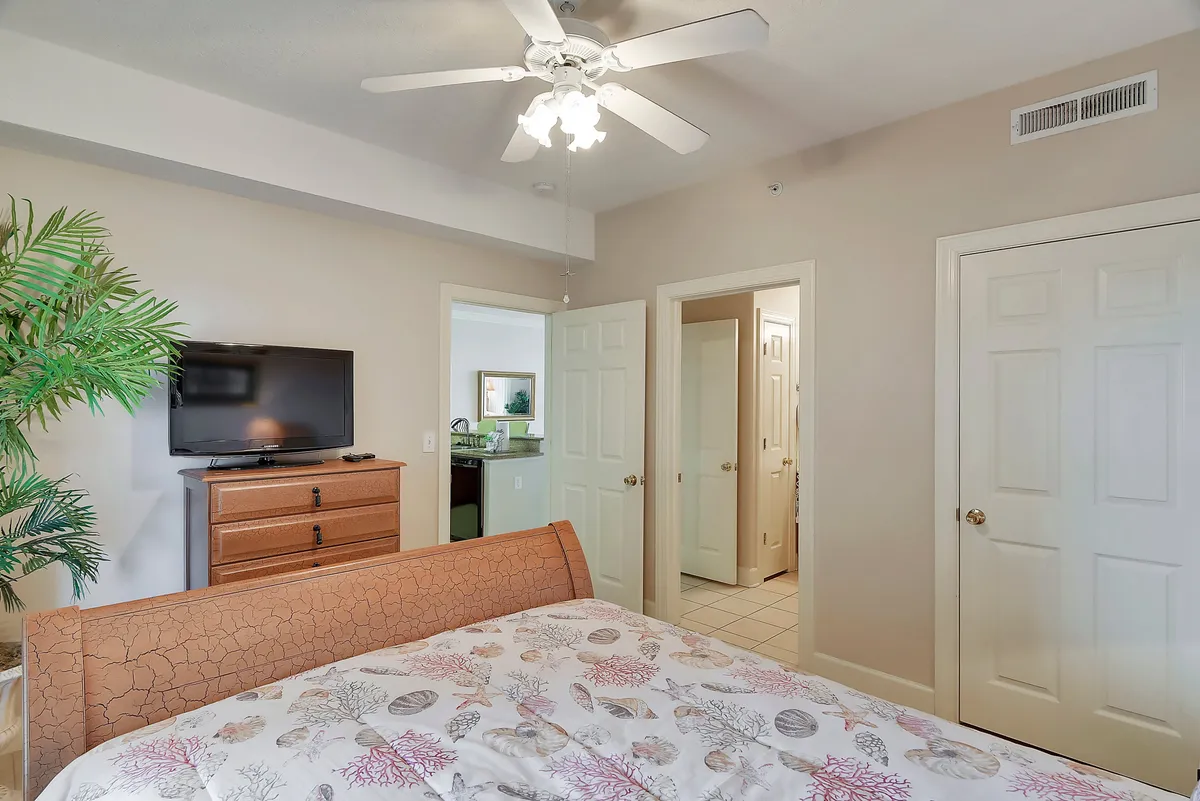 A cozy bedroom with a patterned bedspread, flat screen TV on a wooden dresser, and open doorways leading to both a bathroom and another room. A decorative indoor plant adds a homey touch.