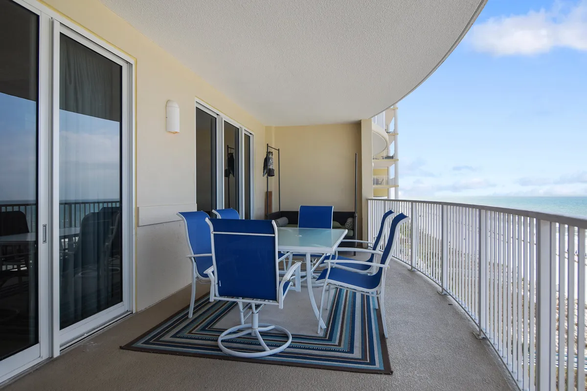 A private, curved balcony furnished with blue cushioned dining chairs and a glass-top table, bordered by a decorative rug and sleek railing that overlooks the expansive ocean. The setting is ideal for al fresco dining, sipping coffee, or unwinding while gazing at the water and sunset.