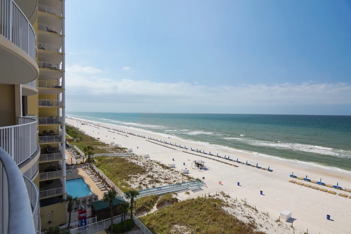 This image presents a wide, inviting sandy beach with arranged umbrellas and chairs, gentle ocean waves rolling onto the shore, and resort balconies overlooking the beach. A small pool area is visible near the beach access, suggesting the ease of moving between sand and poolside relaxation.