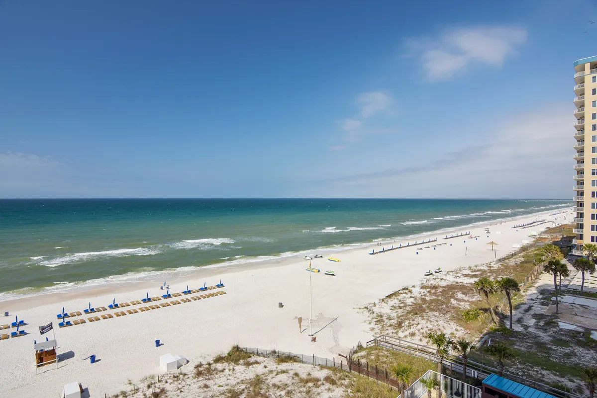 This image features a wide stretch of white sandy beach along the coastline, calm blue-green ocean waves, and neat rows of blue lounge chairs. A tall resort building is visible to the right, connecting the beachfront directly to the accommodations. The scene feels peaceful and inviting, perfect for long walks, sunbathing, and enjoying the ocean view.
