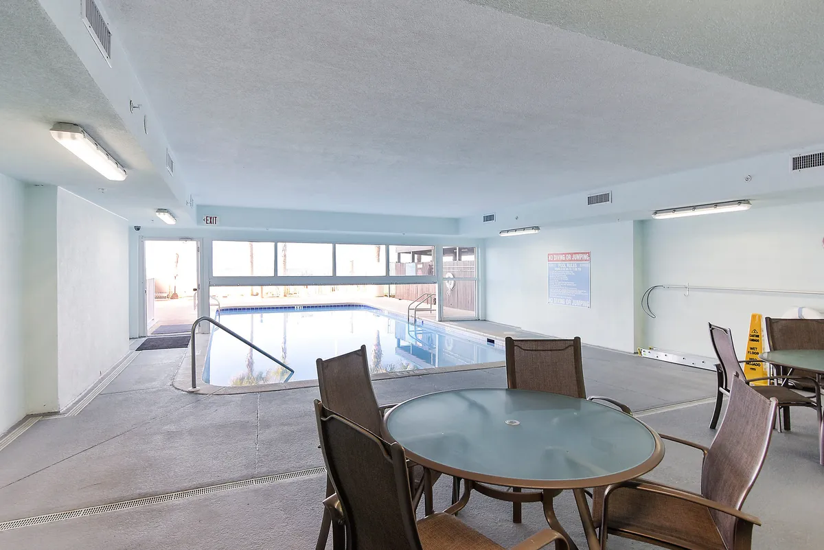 The photo shows an indoor swimming pool surrounded by glass windows that allow natural light to flood the area. Several round glass-top tables with chairs offer a spot for socializing or relaxing between swims, emphasizing the resort's climate-controlled wellness facilities.