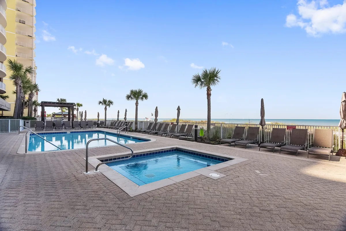 This image captures a swimming pool and hot tub with a row of lounge chairs, all overlooking the beach and ocean. The tranquil scene showcases the pool area as a seamless extension of the beachfront, inviting guests to relax and enjoy both pool and ocean amenities.