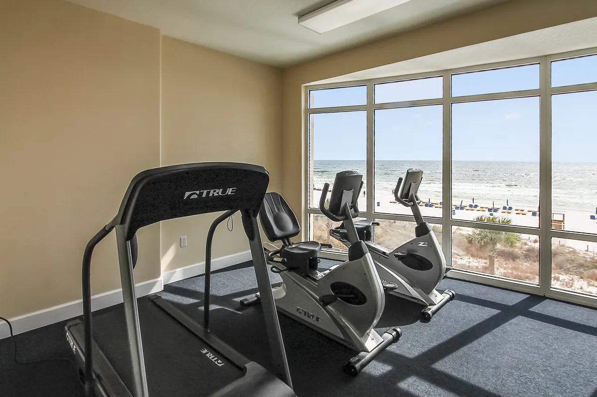 A modern fitness center with a treadmill and stationary bikes faces floor-to-ceiling windows, providing expansive ocean views. The light-filled space is inviting and motivational, highlighting the wellness aspect of the resort.
