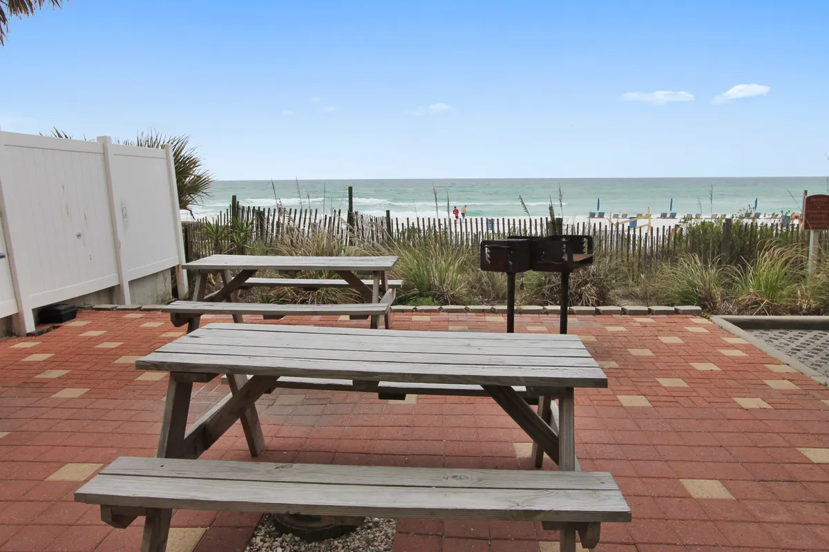 Wooden picnic tables and a charcoal grill are set in an open-air area with a view of the beach and ocean. The scene is casual and welcoming, suggesting opportunities for barbecues, socializing, and outdoor relaxation with the Gulf breezes.