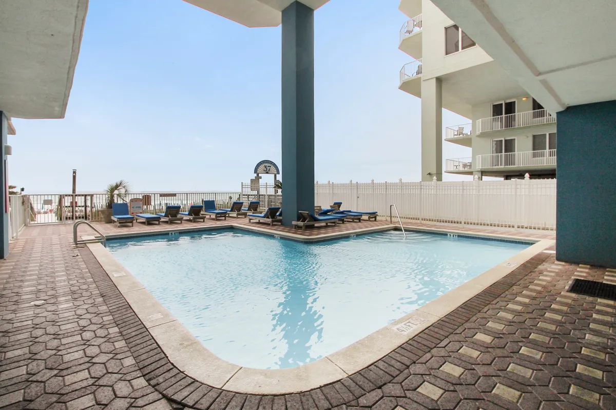This image displays an outdoor swimming pool with sparkling blue water, numerous lounge chairs on the deck, and an ocean view just beyond the pool area. The modern resort architecture frames the scene, and the overall mood is calm and leisurely, ideal for relaxation in all seasons.
