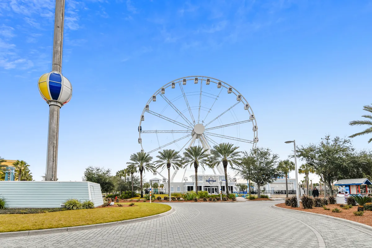 The image showcases a large Ferris wheel (the Sky Wheel), a colorful tower, and a palm-lined road with a curved pathway. The scene is vibrant, lively, and represents one of Panama City Beach's most recognizable attractions, with the promise of community events and seasonal activities nearby.