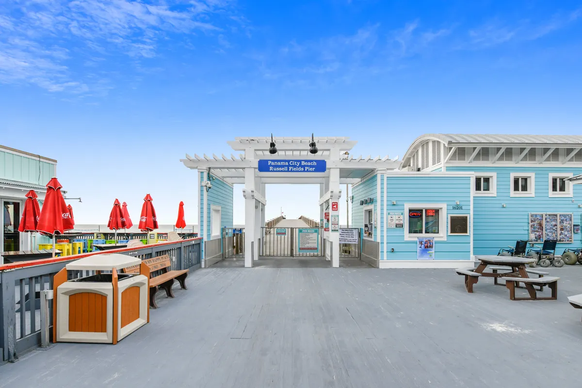 The lively entrance to the Panama City Beach Russell Fields Pier is shown, with blue beachside buildings, white trim, and red umbrellas shading outdoor seating. Picnic tables and benches provide a welcoming, social atmosphere just steps from the sand, highlighting the bustling local dining and entertainment scene.