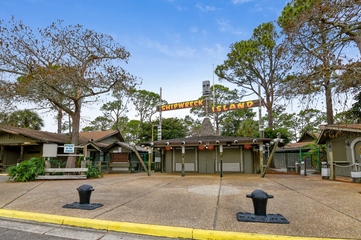The entrance to Shipwreck Island Water Park is featured, with its distinctive sign, rustic wooden structures, and playful pirate-themed decor. The area sets a tone of excitement and anticipation for a fun-filled family outing.